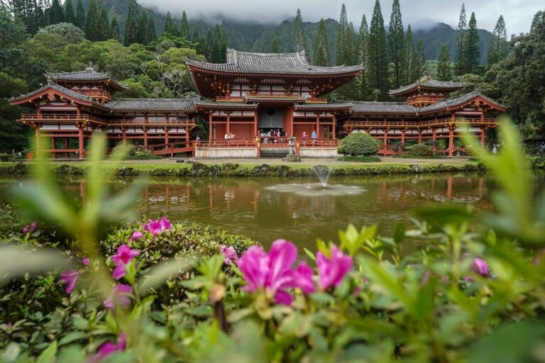 This is a photo of Byodo-In Temple on Oahu... the temple is in the backdrop nestled in greenery while there are pink flowers across a small pond in front of it. How to visit the Byodo-In Temple on Oahu